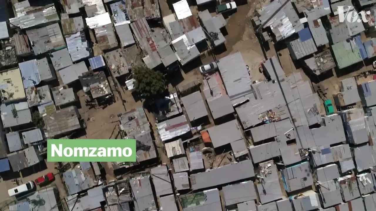 An aerial view of a densely populated area with numerous small structures, labeled 'Nomzamo' with a green overlay. A VOX logo is visible in the upper right corner.