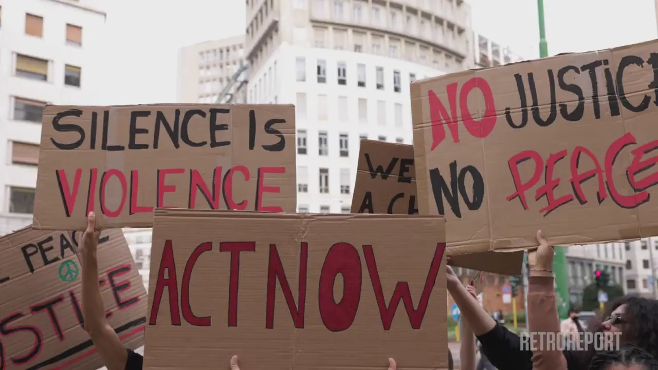 People hold protest signs that read "SILENCE IS VIOLENCE", "ACT NOW", and "NO JUSTICE NO PEACE".