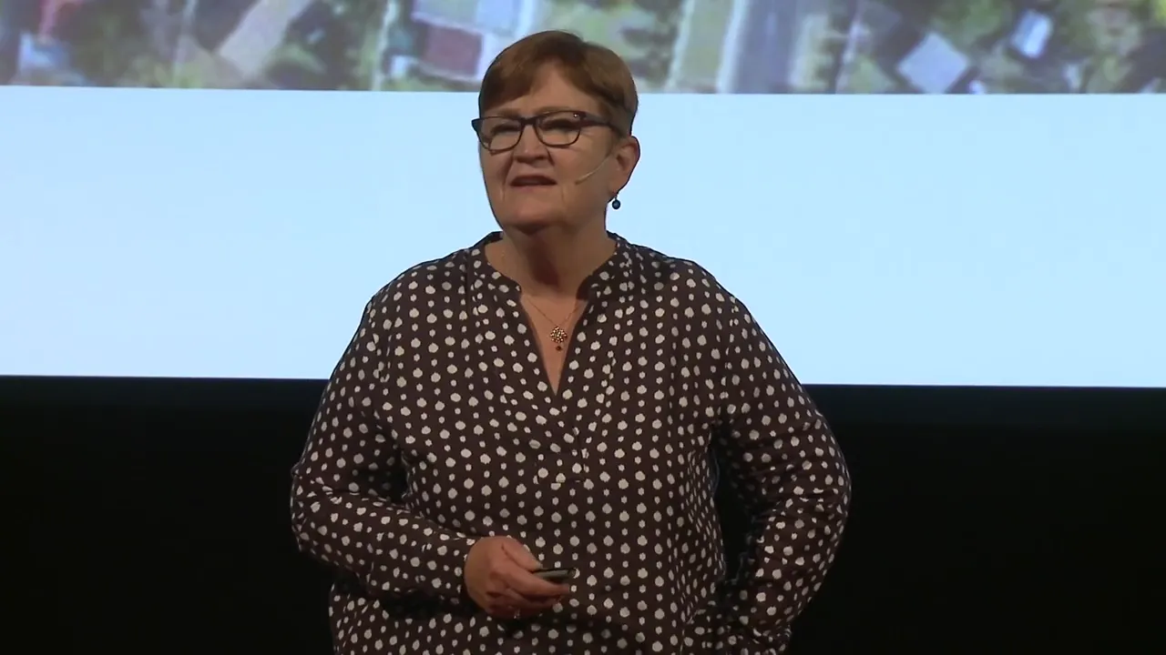 A woman in a polka dot shirt and glasses stands on a stage with a projected image of an aerial view of buildings behind her.