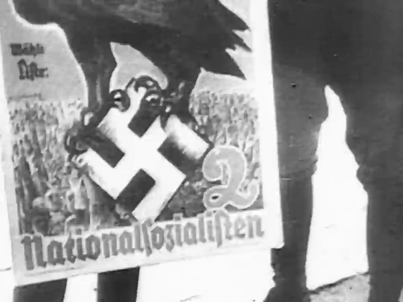 A black and white image shows a poster with a swastika symbol held by a person, with text that appears to be "Nationalsozialisten".